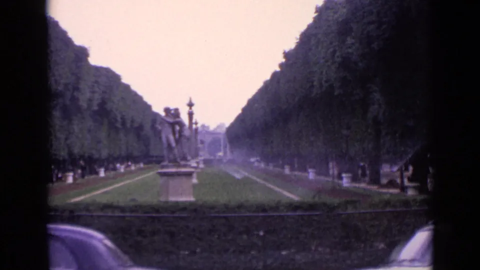 1962 Germany home-movie view of a statue-lined park axis with sprinkler haze, clipped trees, and two classic cars partly visible at the bottom edges.
