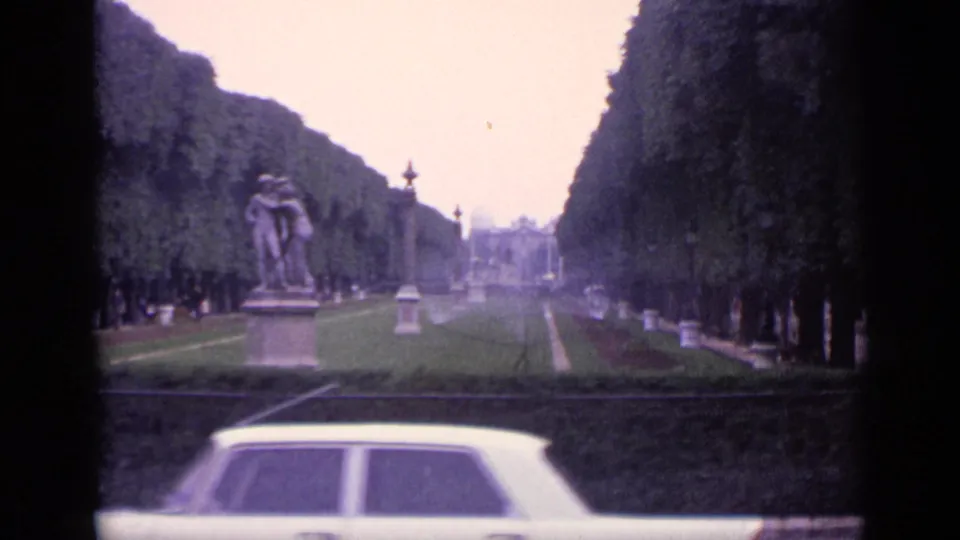 1962 Germany frame showing a formal garden avenue, central pedestal statue, sprinkler mist, distant visitors, and a light-colored classic car in front.
