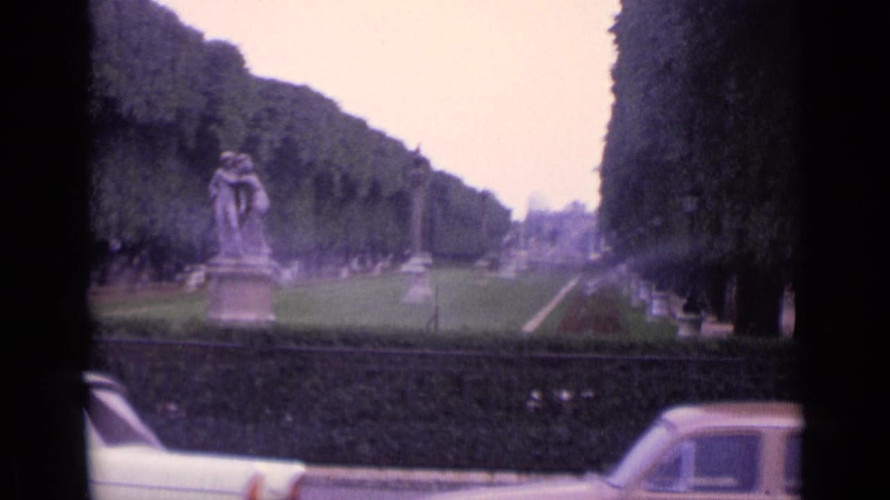 1962 Germany home-movie frame showing a tree-lined boulevard, distant statue, and cars along the lower edge.