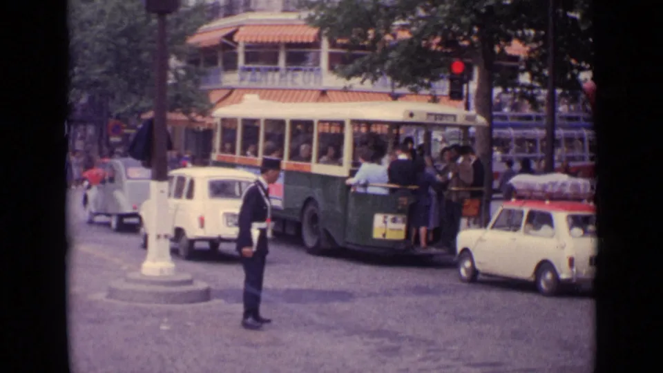 Uniformed traffic officer in 1962 Germany stands near compact cars, pedestrians, and a trolleybus at a city intersection.