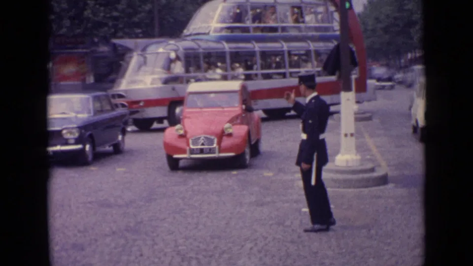 Germany 1962 8mm view of a red compact car moving toward a traffic officer as a glass-roof bus and dark car wait nearby.