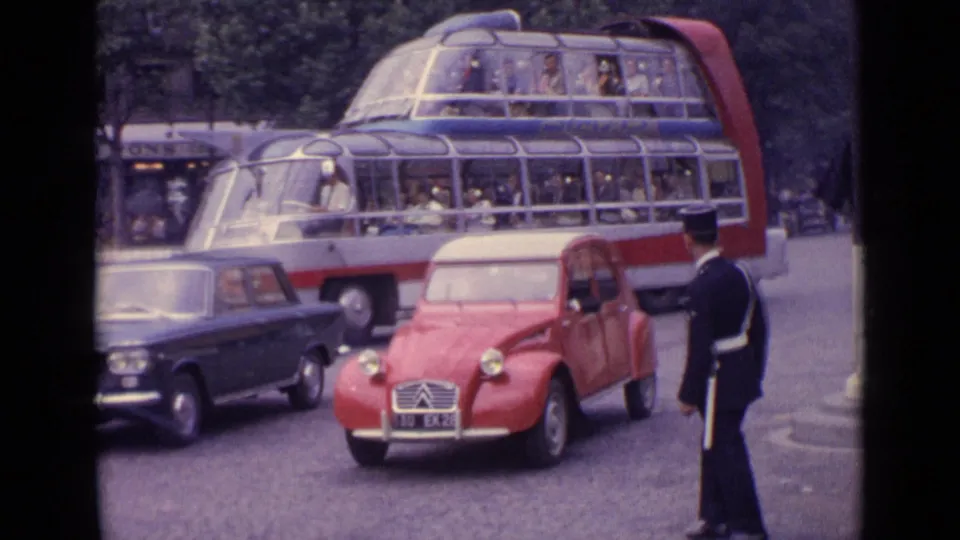 Germany 1962 frame showing a red compact car, dark sedan, traffic officer, and large glass-roof bus carrying passengers.