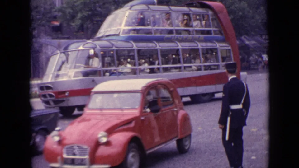 1962 Germany street traffic with a red compact car, a glass-roof double-decker bus full of passengers, and a uniformed traffic officer.
