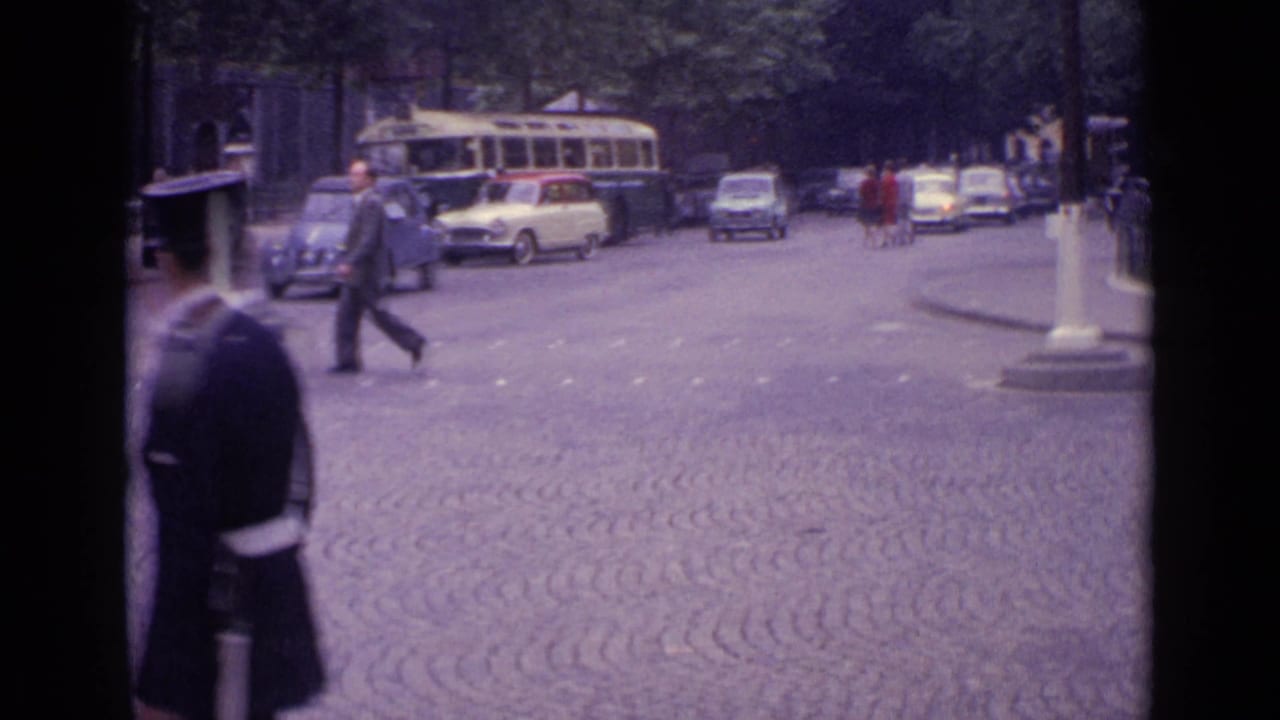 City street with cobblestones, pedestrians, cars, a bus, and a lamp post.