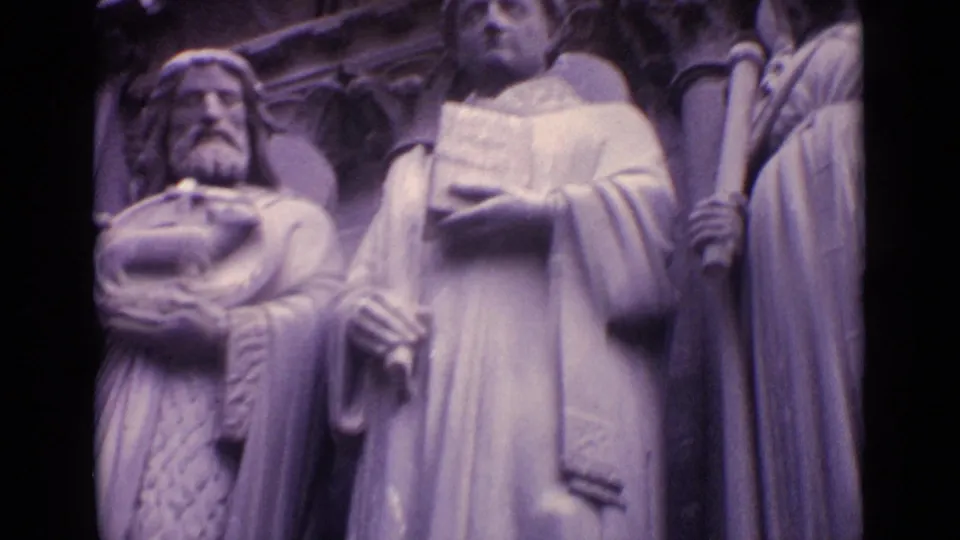 Tight 1962 Germany frame of stone religious facade figures, showing robed figures with a book, staff-like details, and soft 8mm focus.