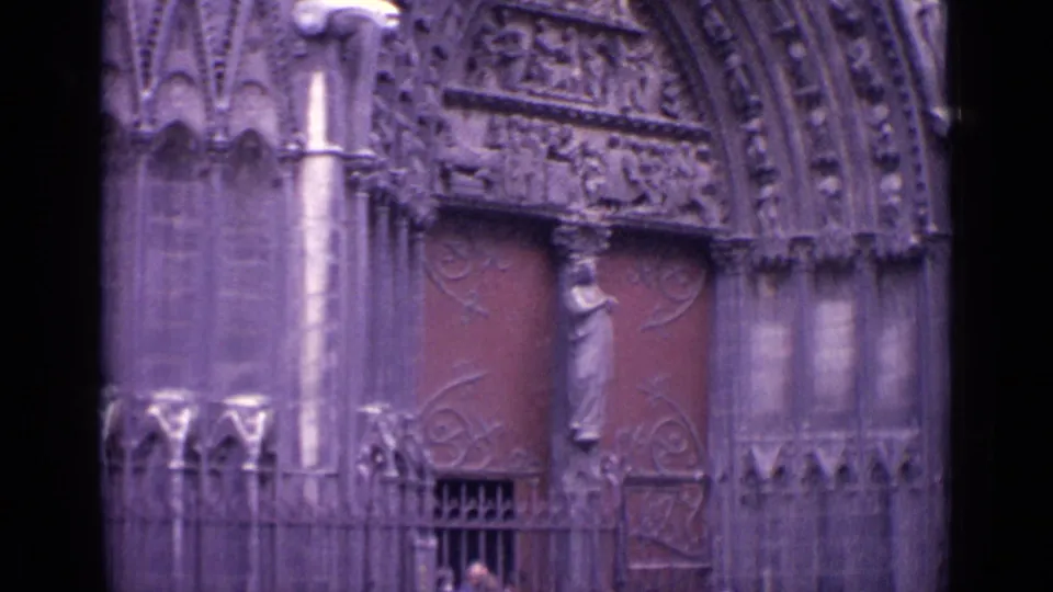 1962 Germany home-movie frame showing Gothic-style church doors, ornate stone carving, iron gatework, and a small visitor near the entry.
