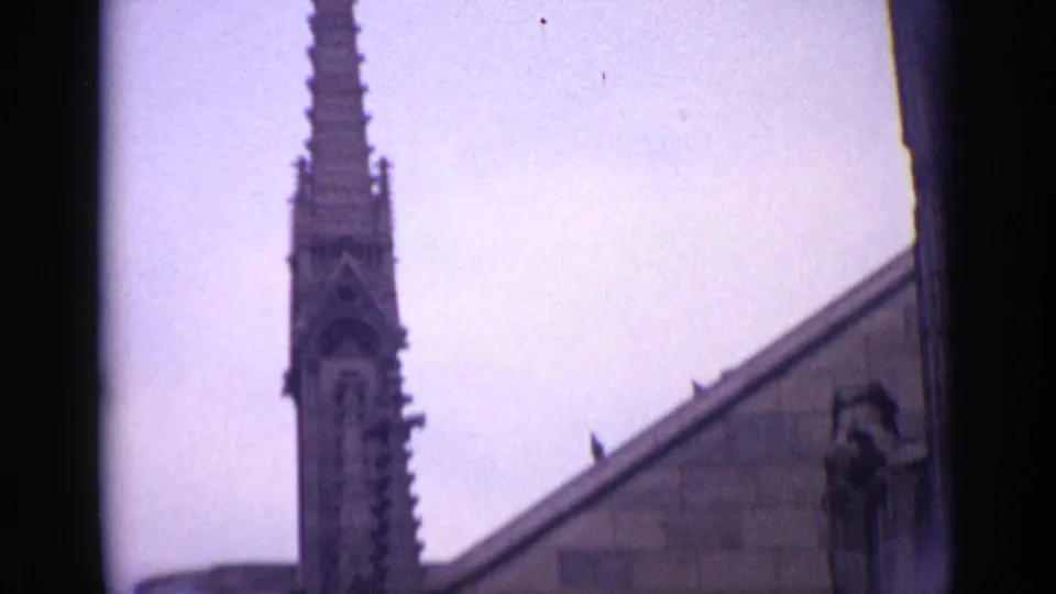 1962 Germany frame with a narrow Gothic steeple above a roofline, filmed from below against a pale sky.