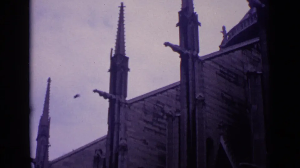 1962 Germany home-movie frame of Gothic church spires, roofline details, and a small bird crossing the overcast sky.