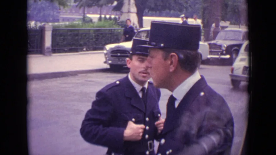 1962 Germany street view with two uniformed men, visible faces, parked mid-century cars, curb, hedge, and trees.