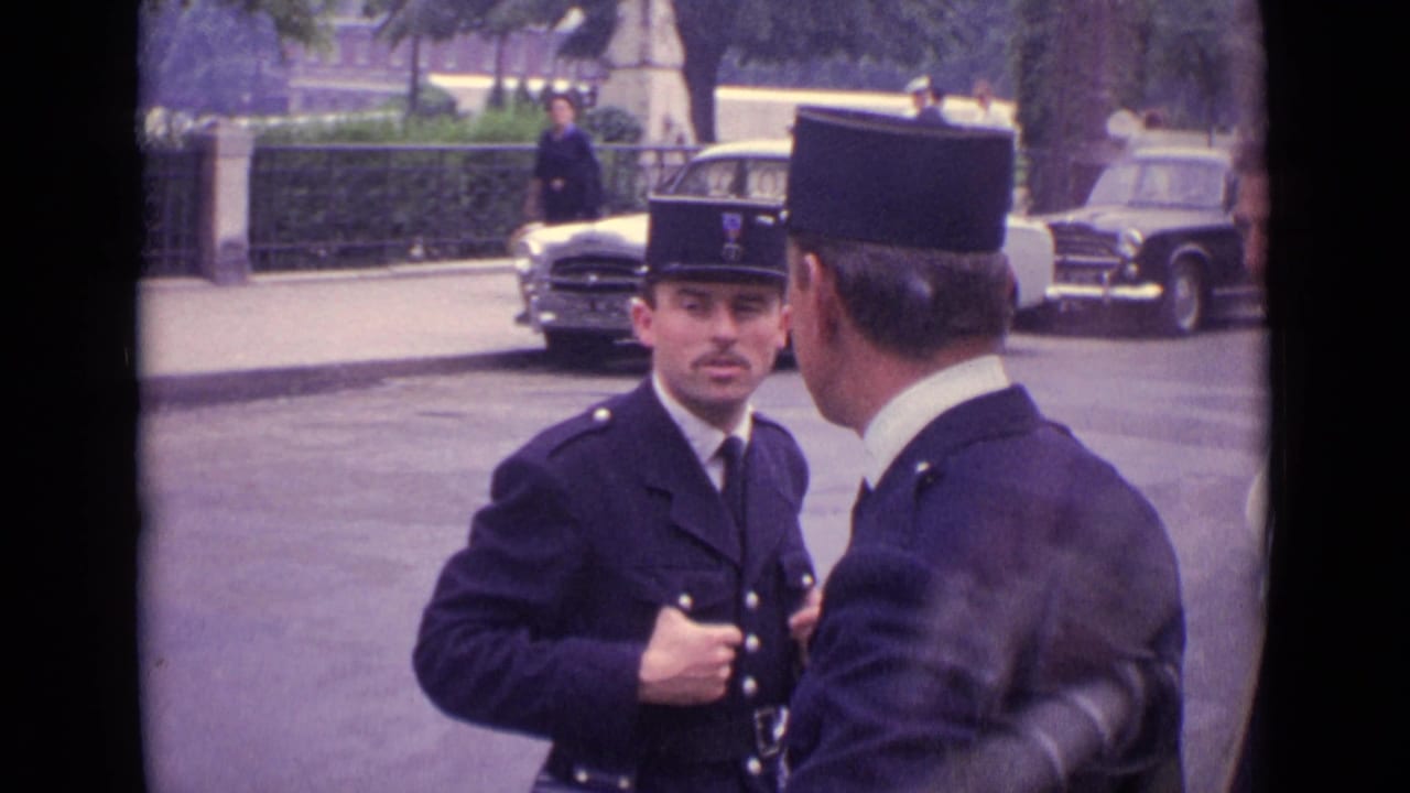 Two uniformed police officers stand on a city street with parked cars behind them.
