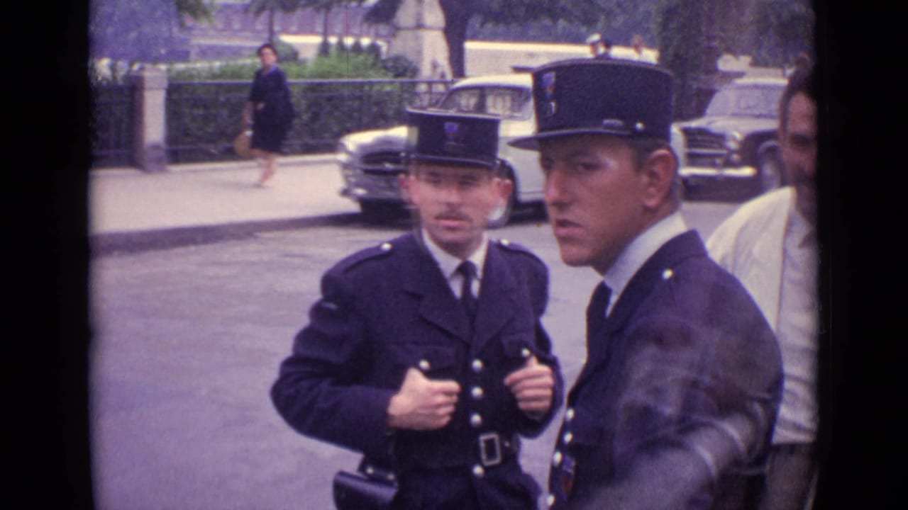 Two German police officers in dark uniforms face the camera area as traffic and parked cars sit behind them.