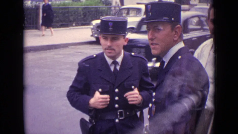 A mustached officer in a dark uniform faces forward while his colleague stands close by on a German street.