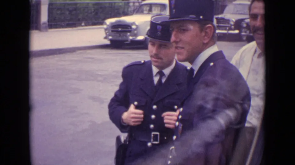Two uniformed officers remain near the curb in 1962 Germany, with parked cars and a partial bystander visible.