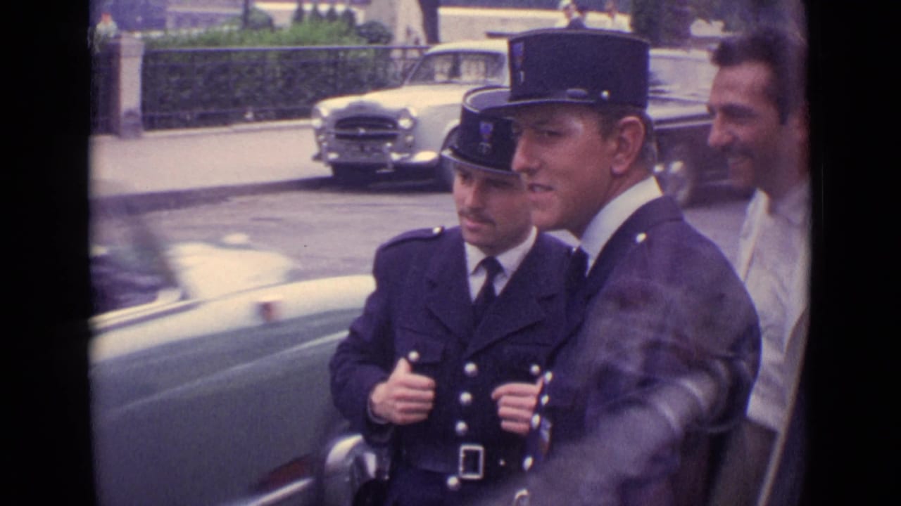 Two uniformed gendarmes walk beside a man in a white jacket on a street with cars behind them.
