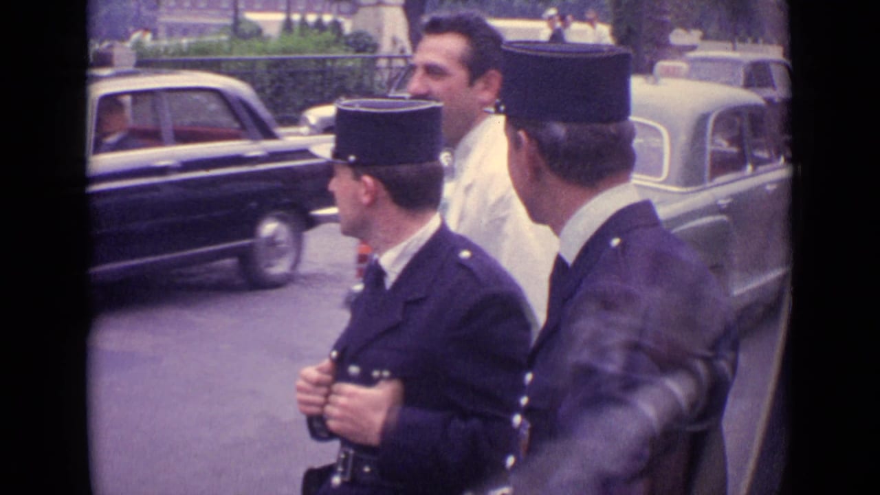 A gendarme looks across the street while another officer and a man in white walk beside moving cars.