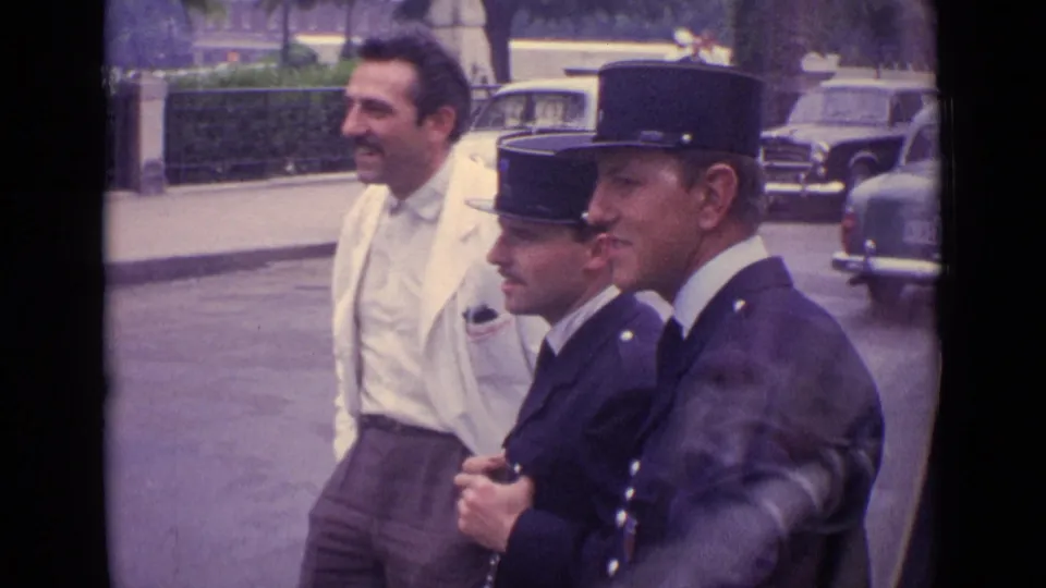 1962 Germany home-movie frame showing a man in a white jacket behind two French gendarmes, with traffic and hedges in the background.