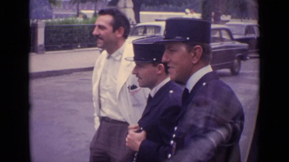 1962 Germany frame showing two French gendarmes in profile with a man in a white jacket, filmed from close range on a street.