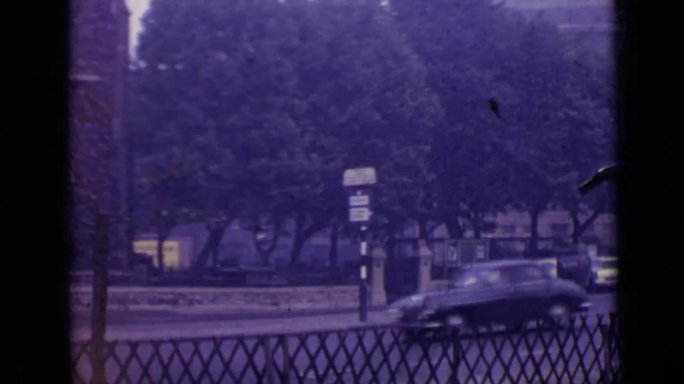 1962 London street view below the skyline, showing roadside trees, railings, signs, and a blurred dark car passing.
