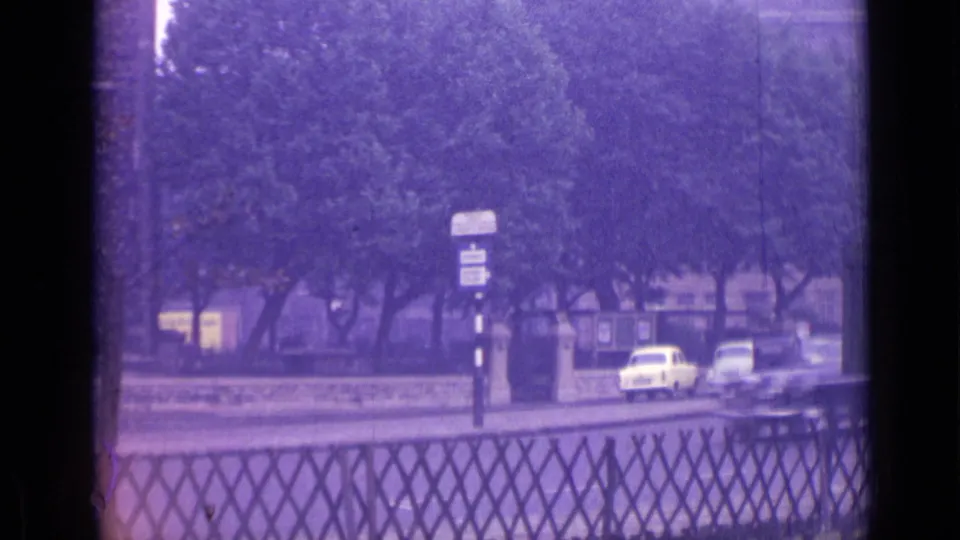 Aged 8mm London street frame with trees, roadside signs, railings, and light-colored cars moving through the foreground.