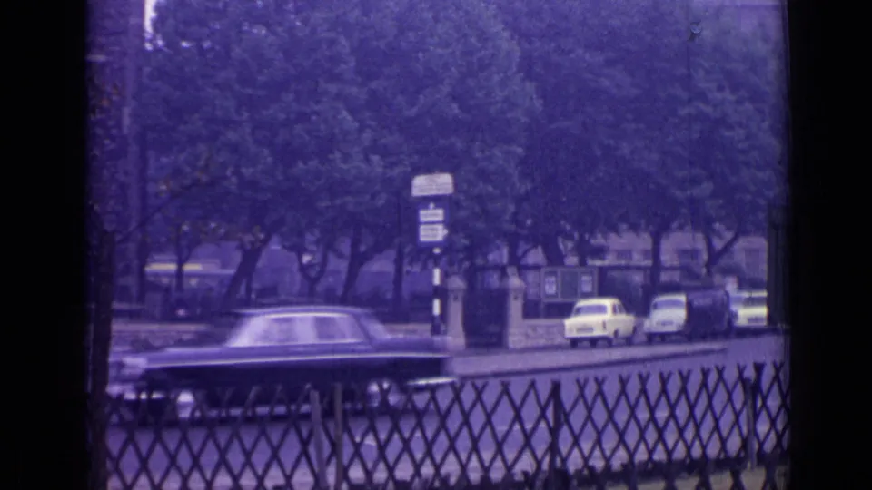 1962 London street view with a dark car passing leafy trees, parked cars, roadside signs, and a diamond-pattern fence.