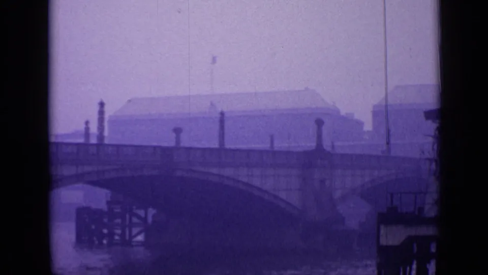 London bridge and river view with a large background building, masonry details, and dark foreground railing in purple-tinted 8mm footage.