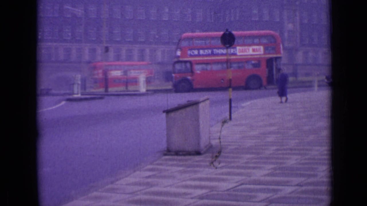 1962 London street view with a red double-decker bus crossing a roundabout, a distant pedestrian, older cars, and a large building facade.
