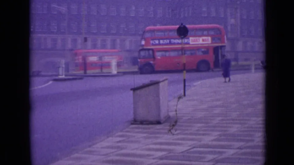 1962 London street view with a red double-decker bus crossing a roundabout, a distant pedestrian, older cars, and a large building facade.