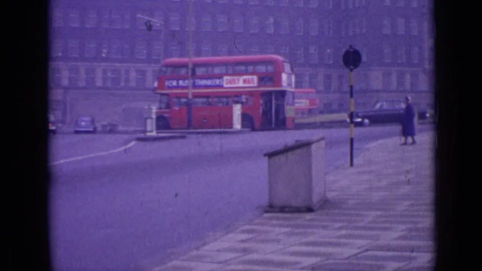 1962 London roundabout scene with a red double-decker bus, older cars, a pedestrian island, and a woman walking along the sidewalk.