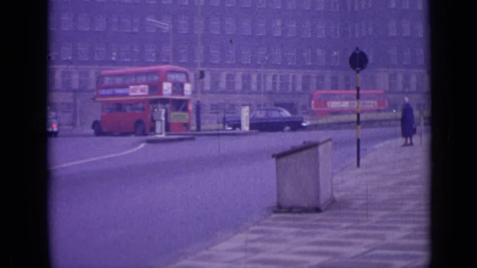 1962 London street frame showing a red double-decker bus leaving the roundabout, a dark car, road posts, and a woman in a long coat.