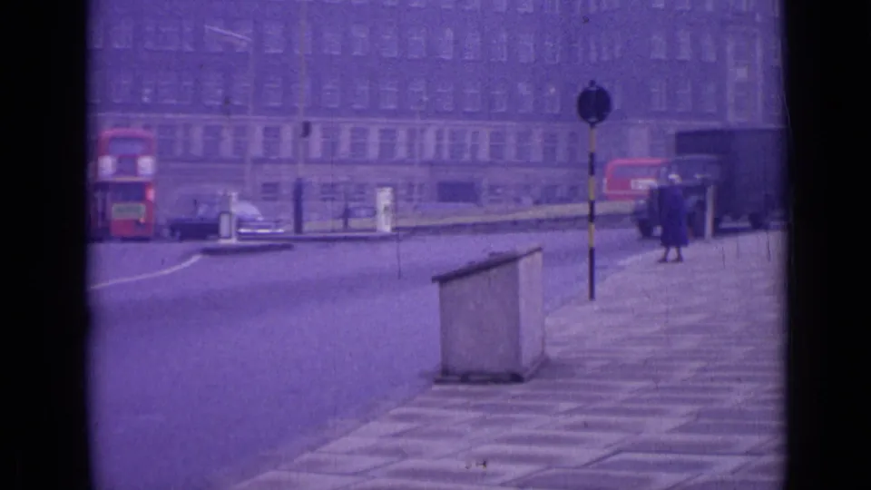 1962 London street view with the rear of a red bus, older cars, a pedestrian post, and a woman walking along the paved sidewalk.
