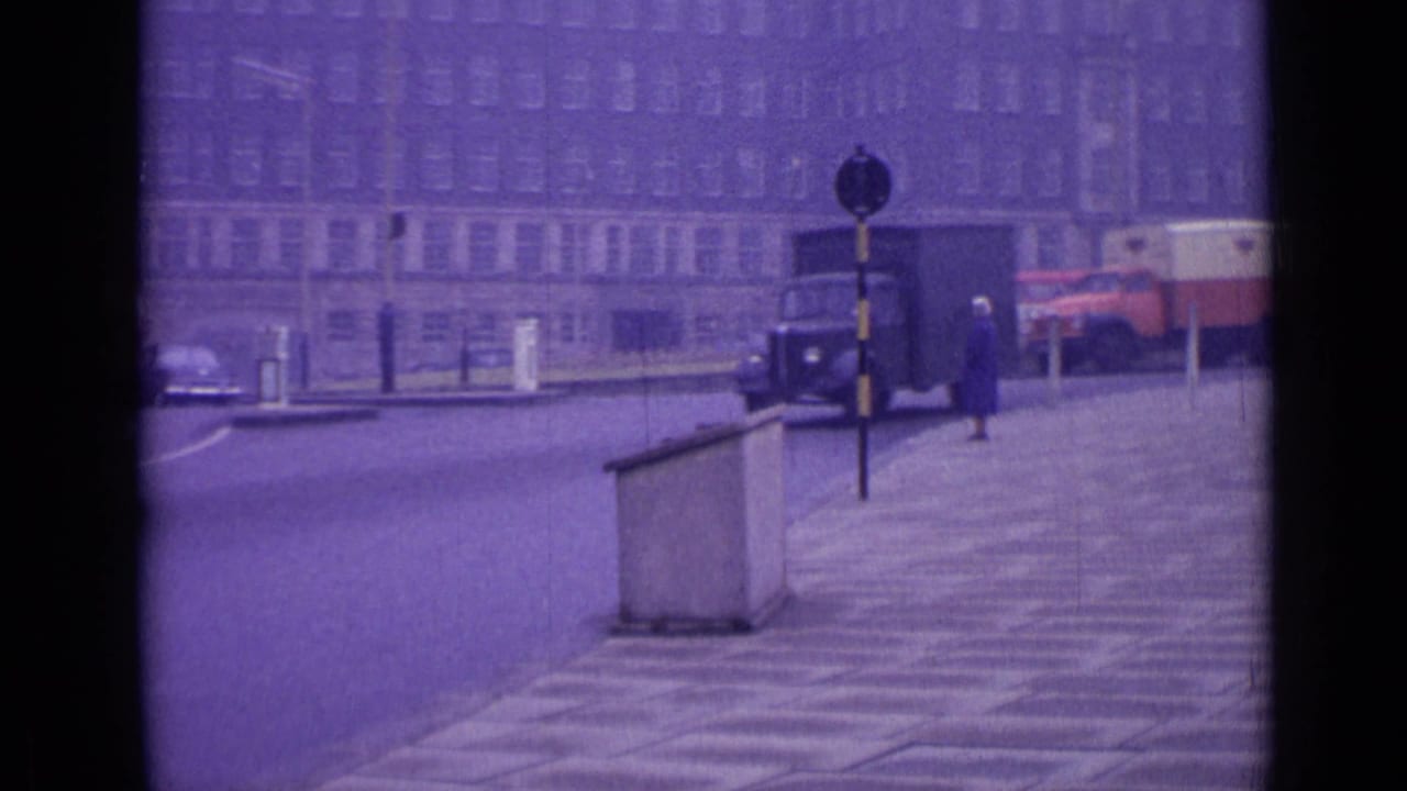 London street with pavement, distant vehicles, a truck, and a pedestrian near a stone barrier.