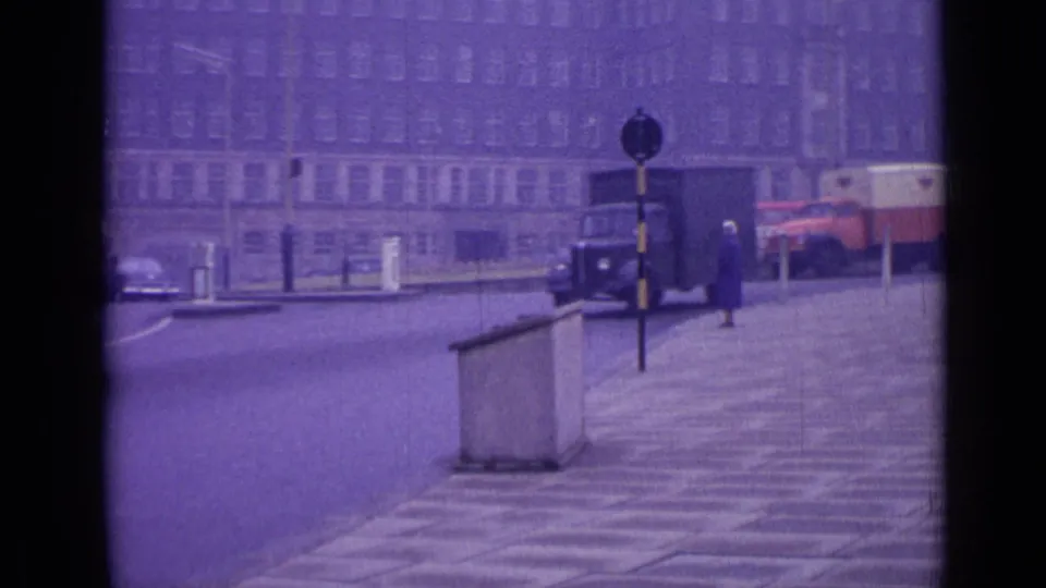 1962 London street view with older vehicles, a boxy truck, pavement, stone barrier, and a distant pedestrian in soft purple 8mm color.