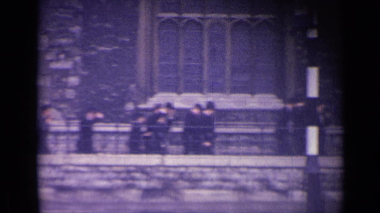 Uniformed boys near a stone wall and railings below Gothic-style windows in London, seen in grainy 1962 home-movie footage.