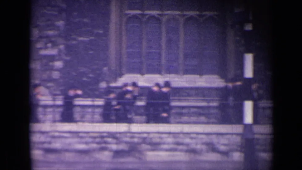 Uniformed boys near a stone wall and railings below Gothic-style windows in London, seen in grainy 1962 home-movie footage.