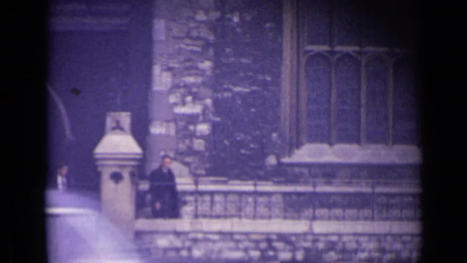 1962 London home-movie frame with a suited man near stone railings and Gothic-style architecture.