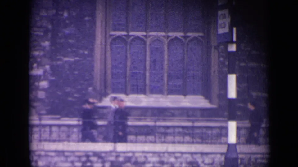 1962 London home-movie frame with figures moving beside a stone railing and historic window.