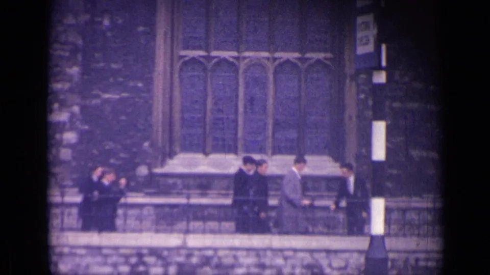 1962 London frame with several suited men near a striped post outside Gothic-style stone architecture.