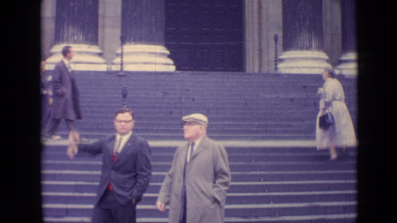 Two men stand on broad stone steps while a woman walks up near large columns.