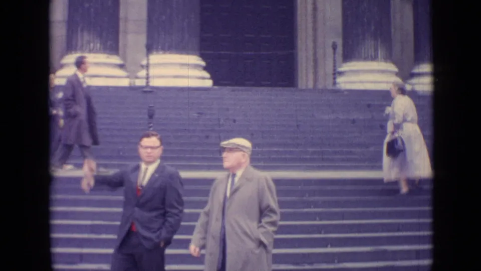 1962 London frame with two men on broad steps, including one in a dark suit, as a woman ascends beside large classical columns.