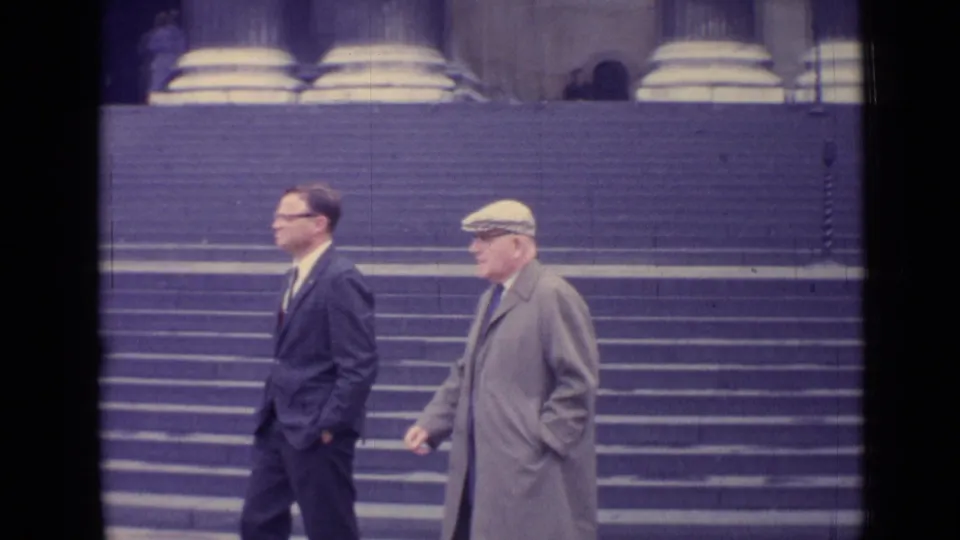 1962 London frame of two men in mid-century dress walking past broad steps and large column bases outside a grand building.