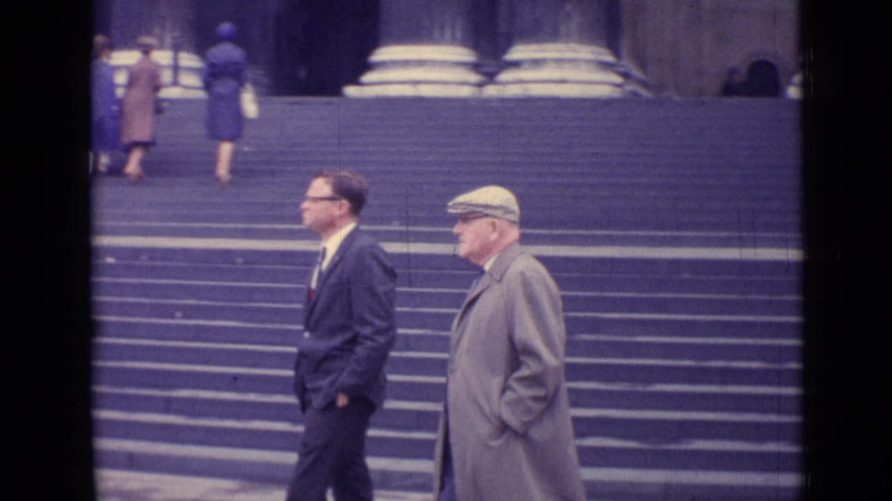 Two men walk past broad stone steps with women near large columns in the background.