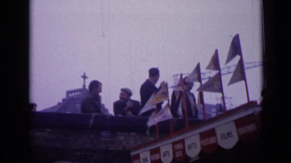 1962 London travel footage shows men beside a souvenir stand with small flags and painted signs for gifts, postcards, and films.