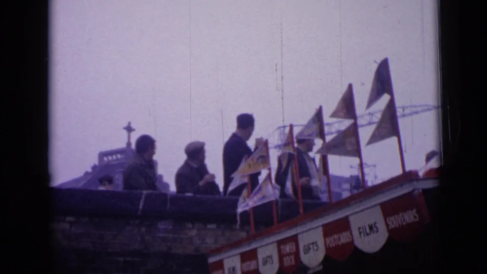 1962 London home-movie frame showing people gathered above street level near small flags and a souvenir stand.