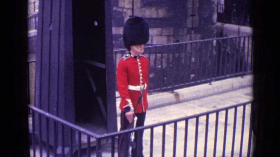 1962 London frame of a ceremonial guard in red uniform and black bearskin hat standing at attention near stone walls.