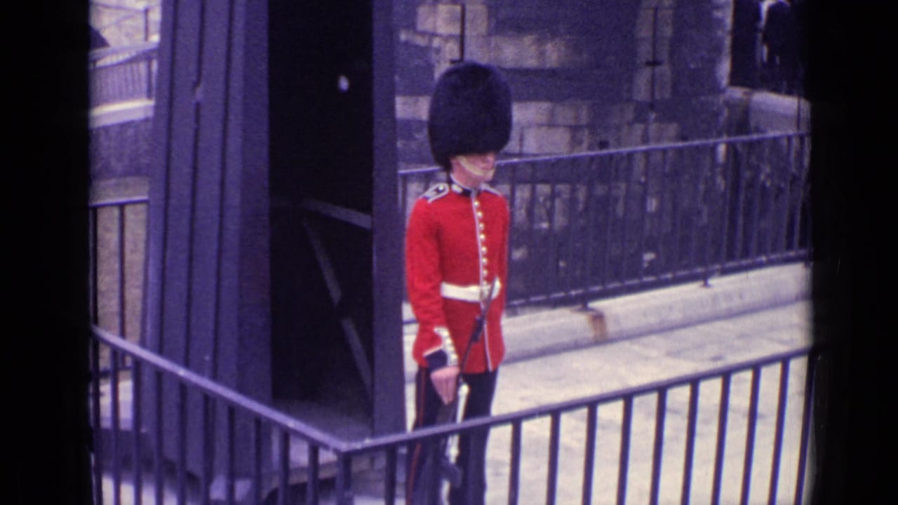 In 1962 London footage, a ceremonial British guard in a red tunic and tall black hat stands near a sentry box and stone wall.