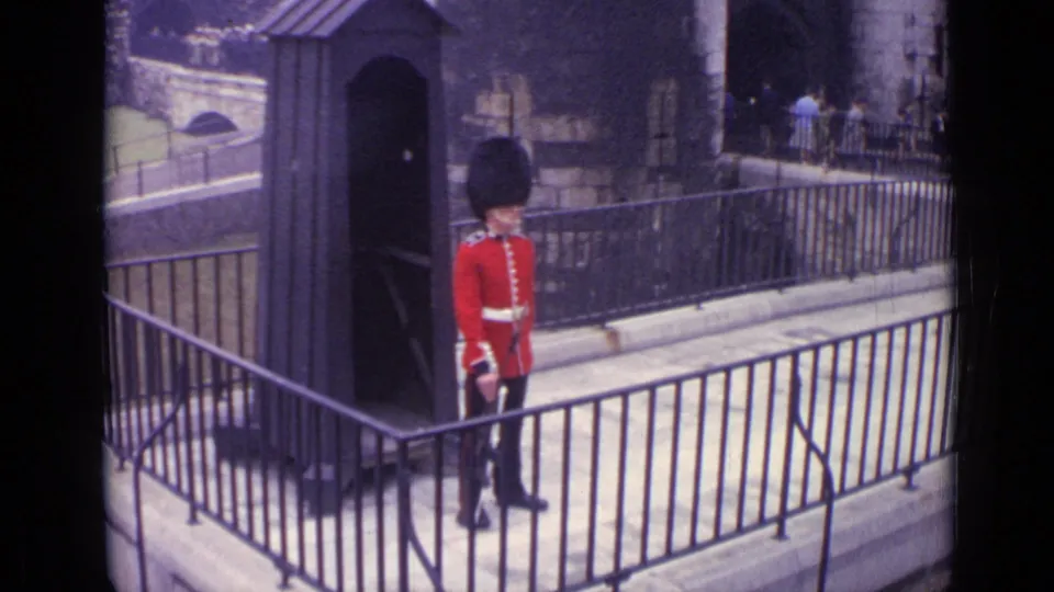 A British ceremonial guard in London stands near a sentry box with a stone fortress wall and metal railings behind him.