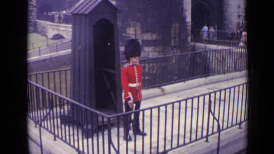 The 1962 London home-movie frame shows a ceremonial guard behind railings, with stone walls and distant visitors in view.