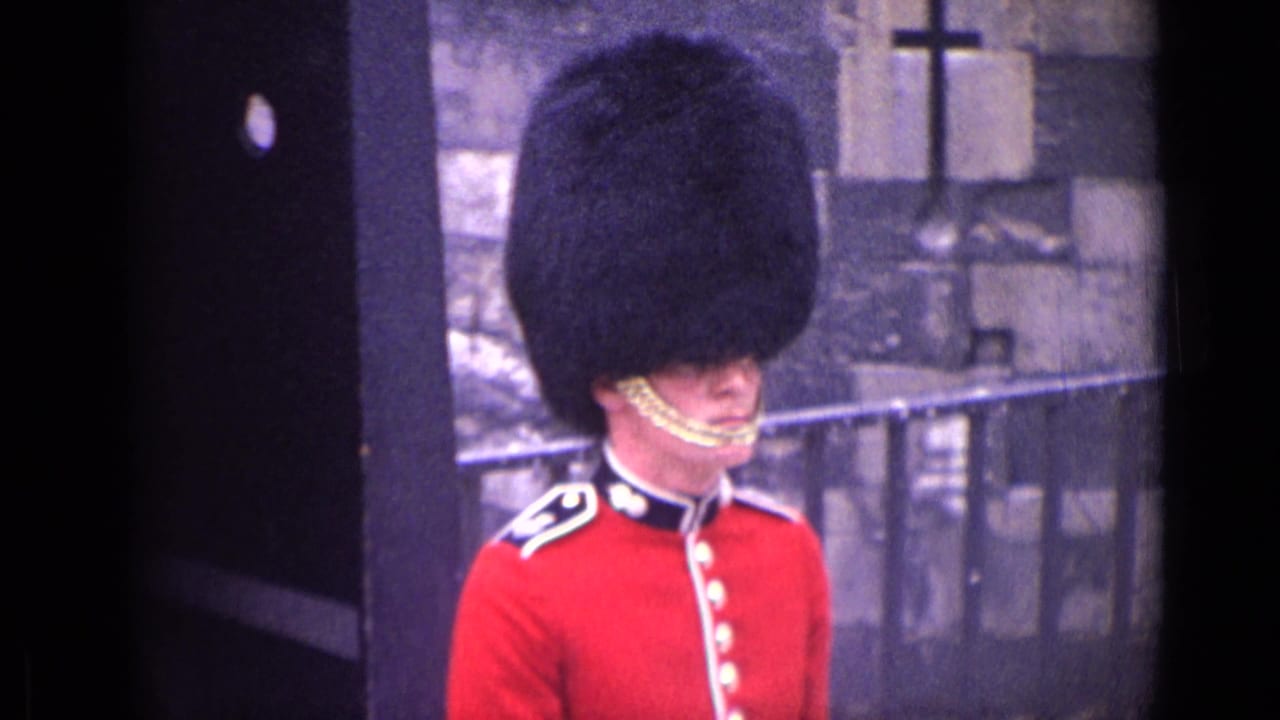 Guard in a red uniform and bearskin hat stands beside a stone building.