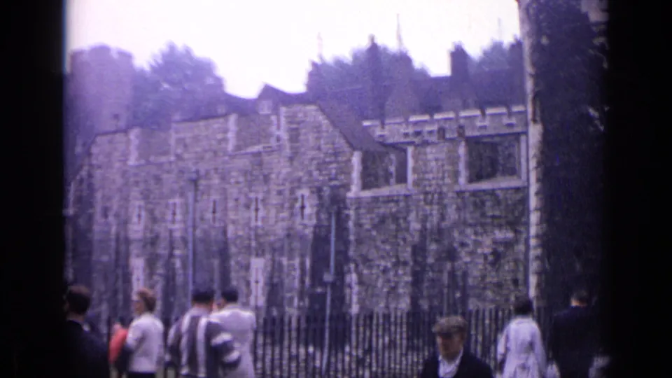 1962 Tower of London scene with tourists near iron railings, stone walls, lampposts and trees on a cloudy day.