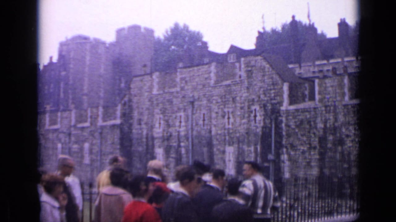 Visitors in 1960s clothing move past the iron fence with the Tower of London's walls behind them.